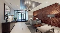 The lobby at 1177 Warburton Ave in Yonkers, NY presents a refined and welcoming first impression through its thoughtfully curated design. The entrance is framed by floor-to-ceiling glass doors that invite natural light deep into the space, complemented by large glass-panel walls that maintain an open, airy feel throughout.

A dramatic woven wood and gold textured accent wall serves as the lobby's centerpiece, adding warmth, depth, and visual interest to the neutral, light-toned palette. Light porcelain tile flooring with subtle geometric inlay patterns runs throughout, providing a clean and polished foundation. Recessed lighting and a statement ring chandelier illuminate the space with a warm, layered glow.

The seating area is elegantly appointed with contemporary upholstered chairs and a sofa, arranged around a glass-and-wood coffee table atop a geometric-patterned area rug. Decorative elements — including sculptural accents, potted greenery, abstract framed artwork, and a geometric-patterned credenza — add personality and sophistication.

A long corridor extends from the lobby, lined with smooth wall finishes and patterned carpeting, providing seamless access to the building's interior. A glass-enclosed lounge area is visible from the main lobby, reinforcing the sense of transparency and connected, modern design. The overall ambiance is polished, upscale, and inviting.