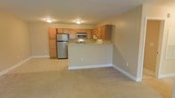 This single-floor space offers an open-concept layout that seamlessly connects the kitchen and living area. The kitchen is equipped with stainless steel appliances, including a full-size refrigerator, microwave, and stove. Medium-toned oak cabinetry lines the walls, providing ample storage space, while a peninsula counter serves as a natural divider between the kitchen and the adjacent living area. The countertops feature a light, neutral finish that complements the warm tones of the cabinetry. Flush-mount ceiling fixtures provide consistent overhead lighting throughout the space. The living area is finished with wall-to-wall carpeting in a neutral beige tone, contributing to a warm and comfortable atmosphere. Tile flooring in the kitchen adds a practical contrast to the carpeted living space. The walls are painted in a soft, neutral beige, creating a cohesive and inviting environment. White baseboards frame the space cleanly, adding a polished finishing touch. The open layout enhances the overall sense of spaciousness, making the area feel bright and functional.
