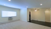 This single-floor space presents an open and airy layout with a clean, neutral aesthetic throughout. The main living area is finished with plush carpeting and features recessed lighting embedded in a smooth ceiling, complemented by a ceiling fan that serves the transition zone toward the kitchen. Crisp white baseboard trim runs along the walls, adding a polished detail against the warm beige wall color.

Natural light enters through a window fitted with horizontal blinds, while a wall-mounted AC unit below provides climate control alongside the baseboard heating system along the perimeter walls. The living space flows seamlessly into an adjacent area with dark hardwood-style flooring, creating a clear visual distinction between zones.

A partial wall separates the living area from what appears to be a kitchen, visible in the background with dark cabinetry and appliances. The open passthrough design maintains a sense of connectivity between spaces. Overall, the layout is functional and straightforward, with practical finishes and essential amenities suited for everyday living on a single level.