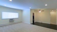 This single-floor space presents an open and airy layout with a clean, neutral aesthetic throughout. The main living area is finished with plush carpeting and features recessed lighting embedded in a smooth ceiling, complemented by a ceiling fan that serves the transition zone toward the kitchen. Crisp white baseboard trim runs along the walls, adding a polished detail against the warm beige wall color.

Natural light enters through a window fitted with horizontal blinds, while a wall-mounted AC unit below provides climate control alongside the baseboard heating system along the perimeter walls. The living space flows seamlessly into an adjacent area with dark hardwood-style flooring, creating a clear visual distinction between zones.

A partial wall separates the living area from what appears to be a kitchen, visible in the background with dark cabinetry and appliances. The open passthrough design maintains a sense of connectivity between spaces. Overall, the layout is functional and straightforward, with practical finishes and essential amenities suited for everyday living on a single level.