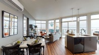 This thoughtfully designed single-floor residence combines contemporary architecture with sweeping waterfront and cityscape views, achieved through an extensive arrangement of floor-to-ceiling windows that flood every room with natural light. Warm hardwood flooring flows consistently throughout, unifying the open-concept layout.

The living area serves as the heart of the home, where oversized windows frame panoramic views of the water and surrounding skyline. The space transitions seamlessly into a dedicated dining area, complete with a marble-topped table and a wall-mounted mini-split system for climate control. A decorative mirror and framed artwork add character to this intimate setting.

The kitchen is sleek and functional, featuring dark flat-front cabinetry, light countertops, black appliances, and additional windows offering water views—making it as visually appealing as it is practical.

Both bedrooms are generously proportioned, each showcasing large windows that capture either waterfront or urban vistas. The primary bedroom features a calming blue accent wall and direct access to natural light on multiple sides.

Two bathrooms serve the residence; one offers a tub-shower combination with modern vanity cabinetry, while the second features a long double-sink vanity with clean, neutral finishes. The overall ambiance is polished, bright, and urban.