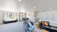 This single-story community clubhouse is thoughtfully designed to serve as a central gathering hub for residents. The main lounge area is generously proportioned, featuring plush blue velvet armchairs, a tufted black leather sofa, and coordinating accent pillows in warm orange and teal tones. Layered area rugs and wooden side tables add texture and warmth to the carpeted floors throughout. Recessed lighting and a textured ceiling contribute to a bright, welcoming atmosphere.

A dedicated resident mailroom wall displays an organized grid of individual metal mailboxes, conveniently located adjacent to the lounge. A wall clock and security camera reflect the practical, community-focused nature of the space. Built-in cabinetry with granite countertops and a large flat-screen television provide an additional media and service area.

The dining zone features a rustic wood table surrounded by modern gray chairs, illuminated by a bold pendant light. A large sliding glass door floods the area with natural light and opens to views of an outdoor pool and lounge deck. An additional room with floor-to-ceiling windows offers pool views and a daybed-style seating bench, creating a relaxed retreat. The overall ambiance is modern, airy, and inviting.