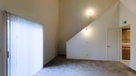 This two-floor space is defined by its striking architectural character, most notably the soaring vaulted ceilings that lend a sense of grandeur and openness to the interior. The dramatic diagonal roofline creates an impressive visual focal point, drawing the eye upward and amplifying the feeling of volume within the space. Soft, neutral wall tones in warm beige run throughout, contributing to a calm and inviting ambiance. Wall-mounted light fixtures provide gentle, ambient illumination that complements the overall warmth of the palette. The floors are finished with plush, light-toned carpeting that adds comfort underfoot and reinforces the space's understated, cozy character. A large vertical blind-covered window or sliding door on one side of the room allows for natural light to filter in while offering privacy and light control. The combination of the angled ceiling, generous vertical space, and soft finishes gives this two-story layout a distinctive and welcoming personality. The open, uncluttered interior provides a flexible canvas well-suited to a variety of furnishing arrangements and uses.