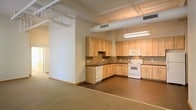 This single-floor space combines an open-concept layout with functional design elements. The kitchen is a standout feature, configured in an L-shape and outfitted with light wood cabinetry that extends to the ceiling, providing ample storage. Dark countertops contrast the cabinetry, while a tile backsplash adds a subtle decorative touch. White appliances, including a dishwasher, electric range, and microwave, are cleanly integrated into the layout. A sink positioned along the lower cabinetry run adds further practicality.

Overhead, exposed white ductwork and piping run along the ceiling, lending an industrial aesthetic to the space. Fluorescent lighting fixtures illuminate the kitchen area effectively. The flooring transitions from hard surface tile in the kitchen zone to carpeting in the adjacent open living area, visually defining each zone without the use of walls.

The living area is spacious and open, offering flexibility in furniture arrangement. A partial column and low partition wall help define the space without enclosing it. The neutral color palette of beige walls and light tones throughout creates a clean, understated ambiance suitable for a variety of uses.