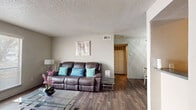 This single-floor residence offers a functional and cohesive layout with consistent wood-look flooring flowing through the main living areas. The living room serves as the heart of the home, featuring a large picture window that fills the space with natural light, a textured ceiling, and an open sightline that connects seamlessly to the dining area. The dining room is a defined, intimate space illuminated by a chandelier overhead, providing an inviting setting for meals and gatherings. Adjacent to the dining area, the galley-style kitchen is well-appointed with white cabinetry, dark countertops, black appliances including a range and dishwasher, and ample storage from floor to ceiling. An entry area provides a welcoming transition into the home, with durable wood-look flooring underfoot. The bedroom is comfortably sized, featuring plush carpeting, a ceiling fan, and a window with blinds that allows natural light to filter in. The bathroom includes a single-sink vanity with cabinetry, a tiled shower-tub combo, and matching wood-look flooring. Throughout the home, neutral wall tones and textured ceilings create a clean, cohesive aesthetic.