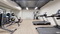 Located at 633 Northcliffe Blvd in Toronto's west end, this compact fitness facility offers a well-equipped workout space on a single level. The room features light wood-look vinyl flooring and crisp white walls, creating a clean and functional environment. Overhead, an exposed dark ceiling with flush LED panel lighting provides bright, even illumination throughout the space.

The gym is equipped with a comprehensive selection of fitness equipment, including a Spirit treadmill, a stationary spin bike, a multi-function cable/pulley machine, an adjustable weight bench, and a full rack of dumbbells in varying weights. Exercise and medicine balls are also available for functional training. A wall-to-wall mirror along one side visually expands the space and allows for proper form monitoring during workouts.

A wall-mounted split air conditioning unit ensures a comfortable climate year-round, while a baseboard heater provides supplemental warmth. Access is provided through a windowed blue door. The overall layout is efficiently organized, maximizing the available floor area and allowing users to move freely between equipment. This facility is well-suited for residents or members seeking a convenient, no-frills space for a complete strength and cardio workout.