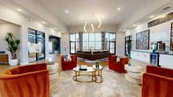 This single-story community clubhouse showcases a thoughtfully designed interior that blends contemporary style with inviting functionality. The centerpiece of the space is a spacious main lounge featuring soaring ceilings, large black-framed floor-to-ceiling windows that flood the room with natural light, and striking globe chandelier pendants. Bold rust-orange velvet accent chairs, a rich brown sectional sofa, and paired circular gold coffee tables are arranged atop a large area rug, creating a warm and sophisticated gathering area. A dedicated seating nook near the entrance offers a tiled fireplace surround with built-in display shelving, two modern swivel chairs, and direct views to the outdoors through expansive glazing. The reception area features a sleek slatted wood front desk, warm hardwood-style flooring, and an open sightline to the adjacent café lounge. The kitchen is well-appointed with gray shaker cabinetry, white quartz countertops, a bold blue hexagon tile backsplash, stainless steel appliances, and warm wood flooring. A café-style lounge area offers mixed seating — high-top tables, round café tables with green velvet chairs, and a tufted blue banquette — set against a textured feature wall. A media lounge rounds out the space with a full wood slat accent wall, a large flat-screen television, and bench seating on patterned carpet, ideal for relaxed viewing.