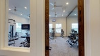 This fitness center offers a comprehensive and inviting environment for residents looking to maintain an active lifestyle. The space is designed with functionality in mind, featuring a well-rounded selection of cardio equipment including multiple treadmills, elliptical machines, and a stationary bike, all positioned along a windowed wall that allows ample natural light to fill the room. A dedicated free weights area includes a full dumbbell rack, catering to strength training needs. A large wall-mounted mirror enhances the sense of space and assists with form during workouts. Recessed lighting and multiple ceiling fans ensure the environment remains comfortable and well-lit throughout the day. Multiple wall-mounted flat-screen televisions are positioned throughout the space, providing entertainment during workouts. The carpeted flooring adds a layer of comfort underfoot, while the neutral-toned walls and white trim create a clean, welcoming aesthetic. The fitness center is accessed through a wood-framed glass door clearly marked with signage, making it easy to locate within the broader property.
