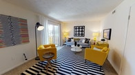 Located at 8470 Towson Blvd. in Miamisburg, Ohio, this single-floor residence offers a thoughtfully designed layout with a consistent yellow, gray, and white color palette throughout. The living room serves as the heart of the home, featuring a sliding glass door that invites natural light and outdoor access. A bold black-and-white striped area rug anchors the space, complemented by vibrant yellow accent chairs and a gray sofa, creating a lively yet cohesive atmosphere. A second sitting room provides a quieter retreat, carpeted and lined with framed artwork, ideal for reading or relaxing. The bedroom is spacious and well-lit by natural light from a large window, featuring plush carpeting, ample dresser storage, and a warm, restful ambiance. An adjacent dressing area with a vanity sink and louvered closet doors adds convenience and functionality. The full bathroom includes a tub-shower combination, a single vanity with generous counter space, and dark cabinetry. The galley-style kitchen is efficiently designed with dark upper and lower cabinets, stone-look countertops, a stainless steel sink, and tile flooring, providing practical workspace for everyday cooking. Throughout the home, textured ceilings, neutral walls, and carpeted floors create a comfortable and inviting environment.