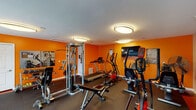 Located at 1739 Tanglewood Ct in Burlington, Kentucky, this single-floor fitness center is a well-appointed facility designed to support a comprehensive workout experience. The space is immediately distinguished by its bold orange accent walls, which energize the environment and are complemented by a clean white ceiling fitted with flush-mount lighting that ensures bright, even illumination throughout.

The main workout area features a wide selection of cardio equipment, including treadmills, elliptical trainers, and upright and recumbent stationary bikes. A wall-mounted flat-screen TV provides entertainment during cardio sessions. For strength training, the facility includes a multi-function cable machine, a weight bench, and a rack of dumbbells, offering users a thorough resistance training setup.

A dedicated functional fitness area provides additional versatility, equipped with a boxing training dummy, stability balls, adjustable dumbbells, and yoga mats stored on a rack — making the space suitable for stretching, core work, and dynamic training. Natural light enters through a set of windows fitted with blinds, offering views of the surrounding outdoor area.

The durable rubber flooring throughout ensures safety and comfort during exercise. Amenities such as a bulletin board, sanitation supplies, and a fire safety station reflect a well-maintained and safety-conscious facility. Two entry doors provide convenient access to the space.