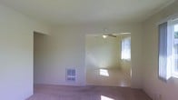 This single-story space presents a straightforward, functional layout with two connected living areas visible from a single vantage point. The main room features wall-to-wall carpet in a neutral tone, smooth painted walls, and a textured ceiling. A wall-mounted HVAC vent near the baseboard provides climate control. An open passage leads into a secondary room, which benefits from a ceiling fan with an integrated light fixture, offering both air circulation and overhead lighting. A window in this adjoining room allows natural light to filter in, casting bright patches across the carpeted floor. The overall color palette is light and neutral throughout, with cream-toned walls and ceilings that contribute to a clean, understated ambiance. The layout feels open due to the wide passage connecting the two areas, allowing light and sightlines to travel freely between rooms. The space is currently unfurnished, making it easy to assess the architectural layout and flow. Overall, the space offers a simple, practical design suited for comfortable everyday living.