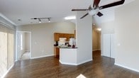 This single-floor residence offers an open-concept layout that seamlessly connects the living and kitchen areas, creating a spacious and functional environment. The rich, dark hardwood flooring extends throughout the main living space, providing a warm and grounded aesthetic that anchors the room.

The kitchen is outfitted with warm oak cabinetry, dark granite-style countertops, and a peninsula breakfast bar that doubles as a casual dining or prep space. White appliances, including a refrigerator and range, complete the kitchen setup, while a skylight above adds a natural brightness to the cooking area.

The living area benefits from vaulted ceilings that amplify the sense of openness and airiness. A ceiling fan with a light fixture is centrally positioned, ensuring both comfort and ambient lighting. Track lighting along the ceiling provides additional directed illumination throughout the space.

Neutral, light-toned walls throughout create a clean and versatile backdrop, well-suited for a variety of interior design styles. A hallway visible from the main area suggests additional private rooms beyond, maintaining a clear separation between communal and private zones. Overall, the space strikes a balance between practicality and comfort, making it an inviting and well-designed home.