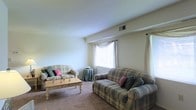 This single-floor residence offers a warm and inviting atmosphere highlighted by a well-appointed living room. The living area is generously sized, featuring soft carpeted flooring that adds a cozy feel underfoot. Two plaid-upholstered sofas serve as the focal point of the seating arrangement, centered around a light wood coffee table that provides both function and charm. The room is brightened by multiple windows dressed with sheer curtains, allowing natural light to filter in and create an airy, open ambiance. A ceiling vent ensures comfortable climate control throughout the space. The neutral-toned walls complement the classic furnishings, contributing to a timeless and welcoming aesthetic. The overall layout is practical and thoughtfully arranged, making efficient use of the available space. The living room's traditional design elements, including the plaid upholstery, soft color palette, and natural light, come together to create a comfortable and functional environment suited for everyday living and relaxed entertaining.