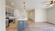 This single-story, 2-bedroom, 2-bathroom home offers a thoughtfully designed layout that maximizes functionality and comfort. The open-concept kitchen and living area serves as the heart of the home, anchored by a striking kitchen island with a gray base and white quartz countertop, fitted with an undermount sink and matte black faucet. A trio of pendant lights with bronze hardware hang above the island, providing both task lighting and visual appeal. The kitchen features gray shaker-style cabinetry, a tasteful tile backsplash, stainless steel appliances including a range, microwave, and refrigerator, and is complemented by recessed lighting throughout. Light-toned luxury vinyl plank flooring flows seamlessly through the main living areas, lending a clean and contemporary aesthetic. The spacious primary bedroom is carpeted for comfort and includes a ceiling fan, ample natural light from a window, and direct access to a private en-suite bathroom. The en-suite features matching gray cabinetry with black hardware and wood-look tile flooring. The secondary bedroom is also carpeted with neutral walls, offering a versatile and cozy space. A dedicated closet provides additional storage, rounding out a practical and well-appointed floor plan ideal for everyday living.
