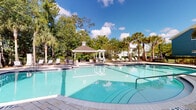 This outdoor pool facility in Jacksonville, Florida offers a resort-style retreat designed for relaxation and recreation. The centerpiece is a generously sized swimming pool with crystal-clear blue water, accessible via broad entry steps with handrails, and framed by a wide paver deck that provides ample space for lounging. A classic white gazebo with a peaked roof serves as a shaded gathering area at the pool's edge, complemented by outdoor seating and tables ideal for socializing.

Surrounding the pool, multiple chaise lounges are thoughtfully arranged on the paver deck, offering abundant sunbathing opportunities. A secondary pool area is also present, similarly outfitted with lounge chairs and bistro-style tables, set against a backdrop of mature trees that provide natural shade and privacy.

The landscaping throughout the space is lush and tropical, featuring tall palm trees, native Florida greenery, and manicured garden beds with colorful ground cover. A gated entry with white railings provides secure access, while the adjacent clubhouse-style building — clad in blue and cream tones with a covered porch and stone columns — adds an inviting architectural presence. The overall ambiance is bright, airy, and quintessentially Floridian, ideal for outdoor leisure year-round.