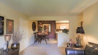 This single-floor residence showcases an open-concept layout that seamlessly connects the living, dining, and kitchen areas into one cohesive space. The living area is anchored by plush carpeting in a neutral beige tone, creating a warm and comfortable foundation throughout. A leather armchair is partially visible in the foreground, hinting at a cozy seating arrangement.

The dining area serves as a focal point, featuring a dark wood dining table with matching cross-back chairs, styled with seasonal floral arrangements in warm orange and red tones. A bold, deep burgundy accent wall provides a dramatic backdrop, complemented by a large framed landscape painting and a decorative circular wall accent, adding visual depth and character.

Adjacent to the dining area, the kitchen is accessible via a pass-through counter, allowing for easy interaction between spaces. It features light wood cabinetry, a white refrigerator, and ample counter space, with a potted plant adding a fresh, natural touch. The overall ambiance is warm and inviting, with neutral walls and soft carpeting tying the spaces together cohesively.