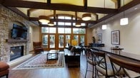 This single-floor space makes a striking impression through its grand architectural design, combining rustic and contemporary elements throughout. Soaring ceilings with bold, dark-stained exposed wood beams create a sense of volume and warmth, while a bank of tall arched and rectangular windows floods the interior with natural light and provides sweeping views of the surrounding landscaped outdoor area.

The main gathering area is anchored by rich hardwood flooring and furnished with a leather sofa and wingback seating arranged around a wooden coffee table, all set atop a traditional patterned area rug. Overhead, a series of large drum pendant lights provide ambient illumination that complements the natural light.

Adjacent to the lounge area, a bar-height counter with cross-back stools offers a more casual seating option, ideal for social interaction. The space transitions seamlessly to the outdoors through a set of French doors leading to a stone patio, enhancing its open, airy character. The overall ambiance is warm and inviting, blending natural materials with thoughtful design to create a versatile and comfortable communal environment.