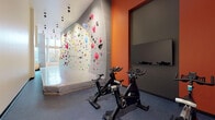 Located at 701 5th Avenue North in Seattle's Lower Queen Anne neighborhood, this single-floor fitness facility offers a dynamic and well-appointed workout environment designed to support a variety of exercise disciplines. The centerpiece of the space is an expansive indoor bouldering wall adorned with a diverse array of colorful climbing holds, spanning a generous length along one side of the room. A padded crash mat runs the base of the climbing wall, ensuring safety during bouldering sessions. The floor transitions from a smooth, light-toned surface near the climbing area to durable rubberized flooring in the cycling zone, where a row of stationary spin bikes is stationed for cardio training. Bold orange accent walls and exposed concrete panels create a striking visual contrast, lending the space an urban, industrial-modern aesthetic. Track lighting mounted along the ceiling illuminates the workout areas effectively. Floor-to-ceiling windows on one end of the facility flood the space with natural light and offer views of the surrounding Seattle neighborhood, contributing to an open, energizing atmosphere. The overall design balances raw, textural materials with functional fitness infrastructure, making this a well-rounded and visually engaging space for climbing enthusiasts and cardio athletes alike.