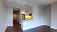 This single-floor space showcases a thoughtfully designed open-concept layout, with the kitchen serving as a central highlight. The kitchen features crisp white cabinetry that extends to the ceiling, providing ample storage while contributing to a clean, modern aesthetic. Stainless steel appliances, including a full-size refrigerator and built-in microwave, add a polished, professional quality to the space.

A peninsula-style counter defines the kitchen boundary, functioning as both a workspace and a casual dining or entertaining surface. Three sleek, metallic pendant lights hang above the peninsula, providing focused task lighting while serving as a contemporary design feature. Under-cabinet lighting further enhances the workspace and adds warmth to the kitchen area.

Rich, dark hardwood flooring runs throughout, offering a striking contrast against the white cabinetry and light grey walls. Recessed ceiling lighting provides consistent illumination across the open living area. A hallway visible in the background suggests additional rooms beyond, contributing to the overall sense of a well-organized and functional floor plan. The overall ambiance is clean, modern, and efficiently designed.