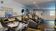 Located at 2922 Chapel Hill Rd in Durham, NC, this dedicated fitness center offers a well-appointed exercise space across a single floor. The room is thoughtfully designed to accommodate a comprehensive range of workout modalities, from cardiovascular training to strength conditioning.

The cardio zone features multiple treadmills, elliptical machines, and a stationary bike, all arranged to maximize floor space and user comfort. A large wall mirror expands the sense of space and allows for form checks during exercise. The strength training area includes a multi-station cable machine, a weight bench, and a full dumbbell rack with varied weights, catering to both beginner and advanced fitness levels.

The interior design is energetic and motivating, with bold blue geometric accent graphics painted across one wall and a series of inspirational fitness quotes—such as "your only limit is YOU" and "the body achieves what the mind believes"—displayed throughout the room. Recessed ceiling lighting provides bright, even illumination, while natural light enters through windows and a glass sliding door, which also offers direct outdoor access. Durable rubber flooring covers the entire space, ensuring safety and functionality. A wall-mounted television and sanitization station add to the room's practical amenities, making this fitness center a complete and inviting environment for daily workouts.