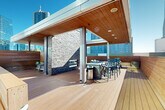 This expansive rooftop terrace is a meticulously designed outdoor living space elevated above a vibrant urban skyline. The deck is finished with warm-toned hardwood planking throughout, creating a cohesive and inviting aesthetic that complements the surrounding cityscape.

A partially covered pavilion anchors the space, featuring a rich wood-paneled ceiling with recessed lighting and supported by sleek structural columns. At the heart of this covered area stands a striking stacked-stone accent wall housing a built-in fireplace and a mounted flat-screen television, providing both warmth and entertainment. Multiple seating arrangements with cushioned wicker chairs and sofas are thoughtfully positioned around this focal point.

Adjacent to the lounge area, a built-in outdoor kitchen and bar counter—also clad in stacked stone—offers bar-height seating for dining and socializing. An open-air sun deck extends beyond the covered pavilion, furnished with hammocks and additional lounge seating, making it ideal for relaxation.

The terrace is enclosed by horizontal wood privacy screens that provide seclusion while maintaining unobstructed panoramic views of the surrounding high-rise skyline. Abundant natural light floods the space throughout the day, enhancing its airy, resort-like ambiance. Overall, this rooftop terrace delivers a sophisticated and functional outdoor retreat in the heart of the city.