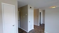 This single-story space presents a clean, functional layout centered around a hallway that connects multiple rooms. The walls are finished in a warm grey tone, complemented by crisp white baseboards and door trim that create a well-defined contrast throughout. Underfoot, dark hardwood-style flooring runs consistently through the space, adding a grounding, rich tone to the overall aesthetic.

The hallway features several white-paneled doors, providing access to adjoining rooms visible in the background. HVAC vents are positioned both on the walls and ceiling, indicating a centralized climate control system integrated throughout. A thermostat is mounted on the wall, reinforcing the practical, utility-focused design of the space.

The ceilings display a textured finish, which is a common and durable surface treatment. Natural light appears limited in this area, with the illumination coming primarily from interior lighting. The layout suggests a straightforward, efficiently designed floor plan with clear circulation paths between spaces. White door frames and clean wall edges contribute to a tidy, well-maintained appearance overall.