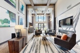 Located at 1050 Blanche St in West Palm Beach, FL, this expansive single-floor community clubhouse offers a generously proportioned gathering space defined by dramatic double-height ceilings and large, grid-paned windows that flood the interior with natural light. The main lounge area anchors the space with a curated arrangement of neutral and teal upholstered seating, a bold abstract area rug, and a sleek linear fireplace framed by an artistic mural accent wall. Designer pendant lighting, including sculptural sunburst and circular ring fixtures, adds visual sophistication overhead. Exposed wood ceiling beams and grey brick column accents provide an industrial-contemporary aesthetic throughout. A casual dining and café zone features round wood tables and modern chairs beneath a striking slatted wood ceiling inset adorned with an artistic Edison bulb chandelier installation. Large glass French doors in the café area open to views of an outdoor pool and palm-lined surroundings, seamlessly connecting the interior to the Florida landscape. A dedicated café counter and work bar offer functional workspace, while a connecting corridor with wood-look plank flooring leads to restroom facilities. Curated contemporary artwork in vivid blues and greens is displayed throughout, enhancing the vibrant, upscale ambiance of this well-appointed communal space.