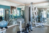Located at 10280 La Hacienda Ave in Fountain Valley, California, this single-level fitness center is designed to provide a comprehensive workout environment. The space is divided into distinct functional zones, each thoughtfully outfitted to support a variety of exercise routines.

The cardio area features treadmills, elliptical trainers, and stationary bikes positioned on protective rubber flooring. Large windows line the walls, flooding the room with natural light and offering views of the surrounding outdoor amenity areas, including what appears to be a pool courtyard. Mounted televisions provide entertainment during workouts, and a wall-mounted air conditioning unit ensures climate comfort.

The strength training zone is visually expansive, thanks to full-length mirror panels that line multiple walls, enhancing the sense of space throughout. This area is equipped with a multi-station cable machine, a leg press, a weight bench, dumbbells in multiple sizes, and stability balls, catering to a range of fitness levels and training styles.

Recessed lighting throughout the ceiling provides consistent, even illumination across both zones. The entryway is clean and functional, featuring a keyed-access glass door, posted facility guidelines, and safety equipment. Warm beige walls paired with teal accent walls create an energetic yet welcoming ambiance throughout the fitness center.