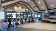 This fitness center offers an impressive and functional workout environment defined by its striking architectural design. The vaulted ceiling with dark exposed wooden beams creates a dramatic sense of height and openness, while recessed lighting and a circular pendant light fixture ensure the space is well-illuminated. Two ceiling fans provide comfortable air circulation throughout the facility.

Large multi-pane windows line the walls on multiple sides, flooding the interior with natural light and offering views of the surrounding outdoor environment. The dark window trim contrasts neatly with the neutral wall tones, lending the space a polished, contemporary aesthetic.

The main workout floor features durable speckled rubberized flooring designed to withstand heavy use. A generous row of elliptical trainers and stair climbers forms the centerpiece of the cardio area, positioned to take advantage of the natural light from the surrounding windows. Additional cardio equipment, including a stationary bike, is visible along the perimeter. Wall-mounted screens and equipment panels provide interactive workout support. The overall ambiance is clean, spacious, and well-organized.
