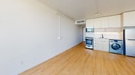 Located at 161 W 600 S in Salt Lake City, Utah, this single-floor unit is situated on an upper level of a modern mid-rise building, offering an open and airy layout with a clean contemporary design throughout. Light wood-tone flooring runs continuously across the main living area, complementing the crisp white walls and creating a bright, cohesive aesthetic.

The living space is defined by a dramatic floor-to-ceiling glass wall with black-framed sliding doors that open to a private balcony. From this elevated vantage point, sweeping views of the Salt Lake City skyline and the Wasatch Mountain range extend across the horizon, making the outdoor space a standout feature. Natural light floods the interior throughout the day through these expansive windows.

The kitchen is efficiently designed with white cabinetry, a stainless steel range, refrigerator, and an integrated washer, providing convenient in-unit laundry. Open shelving with a natural wood and dark metal frame adds a modern organizational element near the kitchen area.

The bathroom features a minimalist, contemporary design with a light wood floating vanity, white countertop, undermount sink, matte black fixtures and hardware, and a large wall mirror, all set against clean white walls and wood-tone flooring. The overall ambiance of the unit is modern, light-filled, and well-appointed.