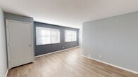 Located at 720 Brittany Square in Grayslake, Illinois, this single-floor unit offers a clean, contemporary interior with a cohesive cool-gray and white color palette throughout. The open living area is finished with light-toned luxury vinyl plank flooring and features dual windows fitted with horizontal blinds, allowing ample natural light to fill the space. The soft gray accent walls contrast nicely with crisp white trim and baseboards, creating a modern yet welcoming ambiance. The layout flows naturally from the living area toward the entry corridor, which includes closet access and ceiling ventilation. The kitchen is efficiently designed in a U-shaped configuration, outfitted with white shaker-style cabinetry, neutral countertops, a stainless steel refrigerator, and a built-in sink with modern fixtures. Track lighting overhead brightens the workspace effectively. The bedroom is carpeted for comfort, painted in the same soft gray tones, and includes a sliding-door closet with built-in shelving and rod storage, along with a window providing natural light. The bathroom features a full tub and shower combination with clean white surround panels and chrome fixtures. Overall, the space presents a well-maintained, move-in-ready interior with consistent finishes and functional design throughout.