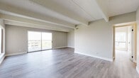This thoughtfully renovated single-floor residence offers a clean, contemporary aesthetic throughout. The expansive main living area is defined by distinctive exposed ceiling beams set against a textured ceiling, all finished in crisp neutral tones. Light gray hardwood-style flooring runs seamlessly throughout, lending a sense of continuity and warmth. Large sliding glass doors open to a private balcony, flooding the living space with abundant natural light and providing open outdoor views of the surrounding cityscape.

The galley-style kitchen is well-appointed with bright white cabinetry accented by matte black hardware, light quartz countertops, a geometric tile backsplash, and a full suite of black stainless steel appliances. A window above the counter brings in additional natural light.

The bedroom is generously sized, featuring the same beamed ceiling detail and a large window that further enhances the airy feel. A hallway leads to the bathroom, which is outfitted with a white vanity with quartz countertop, matte black fixtures, built-in closet storage, and a glass-enclosed shower.

A connecting hallway with textured walls and matching flooring ties all areas of the residence together, completing a cohesive and modern interior design.