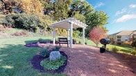 This property is distinguished by its thoughtfully designed outdoor living space, which serves as a central highlight of the grounds. A white pergola anchors the exterior area, providing a defined and shaded retreat. Beneath the pergola sits a classic bench, offering a comfortable spot to relax and take in the surrounding landscape. The pergola's clean, structured lines and latticed roof contribute an architectural element to the outdoor setting.

Surrounding the pergola, the grounds feature carefully maintained garden beds edged with mulch, containing a variety of plantings including low shrubs, ground cover, and flowering plants. A decorative boulder adds a natural focal point to one of the garden beds, enhancing the organic character of the landscape. A charcoal grill positioned nearby suggests the space is well-suited for outdoor entertaining.

The broader grounds are framed by an abundance of mature trees, many displaying autumn foliage in shades of red, orange, and green, lending the setting a vibrant seasonal character. A multi-story brick building is visible in the background, situated amid tall evergreens and additional ornamental trees, all set against a well-maintained lawn. The overall ambiance is peaceful and park-like, with a strong connection to the natural environment.