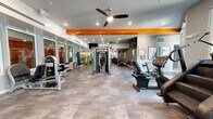 Located at 400 North Rome Avenue in Tampa, FL, this fitness center offers a comprehensive exercise facility designed to support a full range of workout routines. The space is laid out across a single, generously proportioned floor that accommodates multiple training zones without feeling crowded.

The interior is defined by a clean, modern aesthetic with light-toned tile flooring, white walls accented by bold orange structural beams, and recessed lighting complemented by ceiling fans throughout. Large windows along multiple walls flood the space with natural light and provide views to the outdoors, creating an energizing atmosphere.

The cardio zone features treadmills, a stair climber, stationary bikes, and an elliptical machine, all positioned near the windows to take advantage of the natural light. A wall-mounted flat-screen TV keeps users entertained during workouts. The strength training area includes a cable crossover machine, a leg press, and additional weight machines, offering a variety of resistance training options. A dedicated free weights section is outfitted with a full rack of dumbbells, a weight bench, and floor space for functional training. Stability balls and additional accessories are neatly stored and accessible.

Full-length mirrors along one wall visually expand the space and assist with form during exercise. The overall ambiance is bright, functional, and motivating, making this fitness center a well-rounded facility for users of all fitness levels.