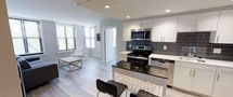 This single-floor space showcases a thoughtfully designed open-concept layout that seamlessly integrates the kitchen and living area. The kitchen is a standout feature, boasting white flat-panel cabinetry, a dark granite or quartz island with bar seating for two, and stainless steel appliances including a range, built-in microwave, and dishwasher. A slate-grey subway tile backsplash adds a modern contrast against the crisp white cabinetry and countertops. Light hardwood-style flooring flows continuously throughout the space, enhancing its clean, cohesive aesthetic. Recessed lighting provides consistent illumination across the open floor plan. The living area benefits from multiple windows fitted with horizontal blinds, allowing natural light to filter in while maintaining privacy. A white interior door leads to an adjacent room, suggesting additional private spaces beyond the main living area. The overall design palette — neutral whites, greys, and blacks — gives the space a contemporary, move-in-ready feel suited for modern living. The open layout maximizes the sense of space, making it feel airy and functional.