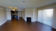 This single-floor residence offers a thoughtfully designed open-concept layout that seamlessly connects the living area and kitchen. The spacious living room is anchored by a classic wood-burning fireplace with a clean white mantel, serving as a natural focal point for the room. Rich, dark hardwood flooring flows throughout the space, adding warmth and a sense of continuity. A ceiling fan provides both comfort and ambient lighting, enhancing the room's functionality. Large vertical blinds cover a full-width sliding door or window, allowing for flexible control of natural light. The open kitchen is well-appointed with dark cabinetry, stainless steel appliances including a range and microwave, and a kitchen island or counter bar, making it an ideal space for both cooking and casual dining. The textured ceiling adds a traditional architectural touch throughout. A hallway visible from the main living area leads to additional rooms, suggesting a practical and well-organized floor plan. Overall, the space presents a clean, neutral palette that offers a versatile and inviting environment.