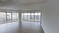 This single-floor space makes a striking first impression with its expansive, open layout and an impressive wall of floor-to-ceiling curved glass windows that wrap generously around the main living area. Situated on an upper floor, the panoramic views stretch across a cityscape of surrounding buildings and a canopy of treetops, filling the interior with an abundance of natural light throughout the day. The clean white walls and smooth, modern ceilings — featuring a circular recessed light fixture and a track rail system — contribute to the space's contemporary character. A sleek, rounded structural column near the window wall adds an architectural focal point while complementing the curved glass facade. Wide-plank hardwood flooring in a warm, medium-toned finish runs throughout the space, grounding the bright, airy atmosphere with a sense of warmth and sophistication. Roller shades on each window panel allow for flexible light control without compromising the open feel. The overall ambiance is modern, luminous, and spacious, making it a versatile canvas well-suited for a variety of uses. The combination of elevated city views, high-quality finishes, and thoughtful architectural detailing defines this space as a refined and distinctive urban environment.
