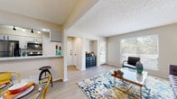 This thoughtfully designed single-floor residence offers a bright and cohesive layout that connects living, dining, and kitchen areas in an open, welcoming flow. The living room serves as the heart of the home, anchored by light wood-style flooring, a textured ceiling, and large windows that invite generous natural light. A patio door provides direct outdoor access, enhancing the airy feel of the space. A smart thermostat is conveniently mounted near the entry, reflecting modern conveniences throughout.

The dining area is defined by a contemporary ring pendant light and features a glass-top dining table surrounded by gold metal chairs, creating a stylish focal point. An adjacent kitchen pass-through connects the two spaces seamlessly. The kitchen is equipped with stainless steel appliances, including a refrigerator and dishwasher, complemented by gray cabinetry and light countertops.

Two bedrooms offer comfortable retreats, each carpeted and fitted with ceiling fans. The bedrooms are well-lit by windows with blinds and teal drapery accents. A hallway connects the bedrooms to a shared bathroom, which features a vanity with dark cabinetry. Throughout, the space is unified by a vibrant, eclectic décor palette that adds personality and warmth to every room.