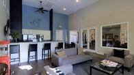 This single-story space showcases an open-concept design anchored by dramatically high ceilings that amplify the sense of spaciousness throughout. The living area is outfitted with a contemporary gray sofa, area rug, and glass-top coffee table, all set atop rich dark hardwood-style flooring. A bold blue accent wall adds a striking visual contrast to the neutral beige surrounding walls, while decorative ironwork wall art contributes a stylish touch.

The kitchen is positioned along one side of the main living area and features white cabinetry, stainless steel appliances, and a granite-topped breakfast bar with dark bar stools — creating a natural divide between the kitchen and the dining space. The dining area accommodates a table with seating for several guests.

A ceiling fan with a black finish is mounted overhead, serving both a functional and aesthetic purpose. French doors with glass panels lead to an adjacent room, allowing natural light to flow freely between spaces and maintaining visual connectivity throughout the floor plan. Recessed lighting throughout enhances the bright, welcoming ambiance of the interior.