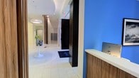This single-floor building lobby offers a welcoming and professionally designed entryway that immediately conveys a modern aesthetic. The space is defined by clean lines, a light-toned tile floor with a subtle circular inlay, and a bright, open layout that promotes a sense of spaciousness and flow. A reception desk finished in warm wood tones sits adjacent to a bold blue accent wall, providing a functional check-in point equipped with a workstation. Sleek black elevator doors serve as a striking architectural contrast against the otherwise light and neutral color palette. A bank of mailboxes lines the far wall, adding a practical residential or mixed-use element to the space. Recessed lighting and a softly illuminated circular ceiling detail contribute to the polished ambiance, while a classic white tulip-style side table with decorative vases adds a touch of refined elegance. The wide, unobstructed corridor extends toward additional access points, offering clear sightlines throughout. Overall, the lobby blends functionality with contemporary design, creating a professional and inviting first impression for residents and visitors alike.