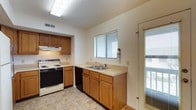 This single-story home offers a practical and functional layout highlighted by a combined kitchen and dining area that serves as the heart of the space. The kitchen is equipped with warm-toned wood cabinetry providing ample upper and lower storage, a dual-basin sink set beneath a window fitted with blinds for adjustable natural light, and a white electric range with an overhead ventilation hood. Black appliances, including a dishwasher, complement the cabinetry throughout. Light-toned countertops run the length of the kitchen, offering generous prep space. A textured ceiling with a recessed fluorescent light fixture illuminates the kitchen area efficiently. The adjoining dining or living area transitions to smooth hardwood flooring, creating a warm contrast to the tile in the kitchen. A stylish pendant light hangs overhead, adding a decorative focal point to the open space. A glazed exterior door with sidelight panel provides direct outdoor access and allows additional natural light to fill the room. The neutral white walls throughout keep the space feeling bright and versatile, ready for personalization.