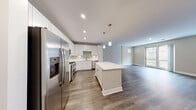 This single-floor unit showcases a thoughtfully designed open-concept layout with clean, contemporary finishes throughout. The kitchen serves as the focal point of the main living area, featuring crisp white shaker cabinetry extending to the ceiling, light quartz countertops, and a soft green subway tile backsplash. A generously sized center island provides additional prep and gathering space, illuminated by elegant gold-accented pendant lights. Stainless steel appliances, including a gas range, built-in microwave, French door refrigerator, and dishwasher, complete the well-equipped kitchen. Warm-toned hardwood-style flooring flows throughout the main areas, transitioning to plush carpet in the bedroom, which features a sliding glass door opening to an outdoor view. An additional flexible room with hardwood-style flooring and a barn-style door offers versatility as a den or second bedroom. Both bathrooms are finished with white subway tile tub-surround enclosures, wood-look flooring, white vanities with quartz countertops, and chrome fixtures. Neutral gray walls and recessed lighting throughout contribute to a bright, modern ambiance.