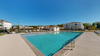 Located at 2200 Cascade Pointe Blvd in Charlotte, NC, this outdoor pool amenity area offers a well-appointed resort-style retreat for residents. The centerpiece is a generously sized swimming pool with crystal-clear blue water, framed by a wide concrete pool deck that provides ample space for lounging and socializing. Numerous chaise lounge chairs are arranged around the perimeter of the deck, offering plenty of spots to relax under the open sky. A dedicated poolside cabana structure with a covered pergola and additional seating provides a shaded gathering area, ideal for outdoor entertaining. A separate utility and restroom building is conveniently positioned at the edge of the pool area. The entire space is enclosed by a black wrought iron fence, ensuring privacy and security. Surrounding views include the multi-story residential buildings of the Cascade Pointe community, as well as mature trees that frame the skyline. The pool area benefits from abundant natural light throughout the day, enhancing its bright, open atmosphere. The overall design is clean and modern, reflecting a community-focused outdoor living experience in a well-maintained residential setting in Charlotte, North Carolina.