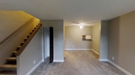 This two-story residence in Willowbrook features two bedrooms and one and a half bathrooms, offering a well-proportioned floor plan designed for comfortable everyday living. The main level showcases a spacious open living area with neutral gray walls, clean white baseboards, and low-pile carpeting that extends throughout, creating a cohesive and understated aesthetic. A ceiling fan serves the main living space, contributing to both function and comfort. A built-in wall shelf adds a practical architectural detail, providing display or storage space without occupying floor area. A closet near the staircase offers convenient main-level storage. The staircase, featuring warm wood-toned treads, connects the two floors and is positioned to one side of the living area, allowing for an open and unobstructed feel in the main living zone. The neutral color palette throughout the main level lends a clean, move-in-ready appearance. The upper floor houses the bedrooms and additional bathroom, keeping sleeping areas separate from the main living spaces. The overall layout is straightforward and functional, making efficient use of the available square footage across both levels.