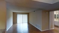 This single-floor residential space offers a clean, functional layout designed for comfortable everyday living. The main living area is a standout feature, showcasing warm hardwood flooring that reflects natural light and adds a sense of warmth to the interior. A full-width sliding glass door, dressed with vertical blinds, serves as the room's focal point, allowing abundant natural light to flood the space and providing direct access to the outdoors. The textured popcorn ceiling adds a classic architectural character to the room, while neutral-toned walls create a versatile backdrop suitable for a variety of interior styles. Practical details such as floor-level heating vents and a ceiling-mounted ventilation register indicate a well-integrated HVAC system throughout. The open layout of the living area lends itself to flexible furniture arrangements, making the space feel expansive and airy. Clean white baseboards frame the perimeter of the room, adding a crisp finishing touch. Overall, the space presents a straightforward and functional residential environment with key comfort features and a bright, welcoming ambiance enhanced by its generous natural light.