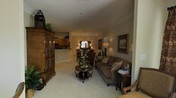 This single-floor home offers a thoughtfully arranged open-concept layout that connects the living, dining, and kitchen areas in a comfortable, flowing arrangement. The living room serves as the centerpiece, anchored by a substantial wooden armoire with decorative glass-panel doors that adds a classic, traditional character to the space. A plush, earth-toned sofa adorned with patterned throw pillows sits opposite a round, ornate coffee table topped with a decorative floral arrangement, lending warmth and elegance to the room. Neutral-toned carpeting extends throughout, contributing to the cozy and cohesive atmosphere. Framed artwork on the walls and a tall, dark decorative accent piece complement the traditional aesthetic. Beyond the living area, a dining space is illuminated by a classic chandelier, creating an inviting transition toward the kitchen, which features warm wood cabinetry visible in the background. Additional ambient lighting from table lamps enhances the warm, welcoming tone throughout. The textured ceiling and neutral wall tones tie the entire space together, resulting in a harmonious, traditionally styled interior suited for comfortable everyday living.