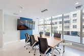 Located at 120 N Compass Way in Dania Beach, Florida, this single-floor professional space offers a polished, contemporary environment designed for productivity and collaboration. The layout is divided into two distinct yet seamlessly connected areas: a formal conference room and an open lounge/reception corridor.

The conference room is the centerpiece of the space, featuring a large rectangular wood-top table surrounded by ergonomic black and blue executive chairs. A wall-mounted flat-screen display is set against a bold blue accent wall, serving as a focal point for presentations. A sleek linear pendant light hangs above the table, providing focused illumination. Expansive floor-to-ceiling windows wrap around the room, flooding the interior with abundant natural light and offering open views of the surrounding Dania Beach community.

The adjacent lounge and corridor area provides a welcoming transitional space, finished with large-format light-toned floor tiles that run throughout. Plush navy lounge chairs and a warm wood side table create a comfortable seating arrangement, while curated abstract artwork adorns the white walls, adding a refined artistic touch. Full-height glass partitions along one side maintain visual openness while defining the space. The overall design aesthetic is clean, modern, and professional, making this space well-suited for business meetings, client receptions, or collaborative work sessions.