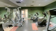 This fitness center is designed to support a variety of exercise needs within a single, efficiently organized space. The room features sage green accent walls paired with a dropped ceiling fitted with recessed lighting, creating a functional and energizing atmosphere. The flooring consists of interlocking rubber tiles in grey and coral, providing both durability and cushioning underfoot — well-suited for active use. A multi-station cable and weight machine anchors the strength training area along one wall, complemented by a wall-mounted mirror that enhances the sense of space. The cardio section hosts an upright stationary bike and a stair climber or elliptical-style machine, offering options for cardiovascular fitness. A frosted-glass door leads to an adjacent room or corridor, suggesting additional amenities nearby. Wall-mounted fixtures and ventilation grilles are thoughtfully integrated throughout. The overall layout is clean and purposeful, making efficient use of the available floor space while maintaining clear pathways between equipment for safe and comfortable use.