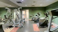 This fitness center is designed to support a variety of exercise needs within a single, efficiently organized space. The room features sage green accent walls paired with a dropped ceiling fitted with recessed lighting, creating a functional and energizing atmosphere. The flooring consists of interlocking rubber tiles in grey and coral, providing both durability and cushioning underfoot — well-suited for active use. A multi-station cable and weight machine anchors the strength training area along one wall, complemented by a wall-mounted mirror that enhances the sense of space. The cardio section hosts an upright stationary bike and a stair climber or elliptical-style machine, offering options for cardiovascular fitness. A frosted-glass door leads to an adjacent room or corridor, suggesting additional amenities nearby. Wall-mounted fixtures and ventilation grilles are thoughtfully integrated throughout. The overall layout is clean and purposeful, making efficient use of the available floor space while maintaining clear pathways between equipment for safe and comfortable use.