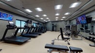 The Village fitness center is a thoughtfully designed, single-floor facility built to support a comprehensive workout experience. The space opens into a welcoming reception area, complete with branded signage, dark wood cabinetry, a digital display screen, and a water station — setting a professional and polished tone from the moment of entry.

The main workout floor is generously proportioned, featuring durable speckled flooring and a dropped tile ceiling fitted with an array of recessed and panel lighting that ensures consistent, bright illumination throughout. A row of treadmills and elliptical machines lines the perimeter, positioned near large windows that offer views of the surrounding outdoor landscape, allowing natural light to enhance the cardio experience. Multiple wall-mounted televisions are positioned throughout for entertainment during workouts.

The strength training area is well-appointed with a robust selection of cable machines, weight benches, and a full dumbbell rack, complemented by floor-to-ceiling mirrors that create a sense of expansiveness. A dedicated Kids Zone area adds notable family-friendly functionality, featuring colorful seating and cheerful wall graphics, providing a safe space for children. The overall aesthetic is modern and utilitarian, with a dark gray color palette and clean lines defining the facility's professional atmosphere.